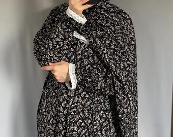 May include: A black prayer dress featuring a small white floral pattern throughout. The garment includes a matching head covering and long sleeves finished with white lace trim at the cuffs. The fabric drapes loosely over the body.