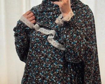 May include: A floral print prayer dress in black, teal, and white. The dress features a matching head covering and white lace trim on the sleeves and neckline. The person is holding a black phone.