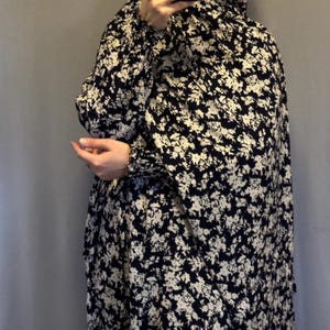 Può includere: Un abito lungo e fluido con un motivo floreale in bianco e nero. L'outfit include un copricapo abbinato. La persona tiene un telefono davanti al viso.