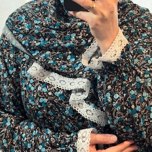 May include: A black garment with a teal floral pattern and white lace trim. The garment has a hood and white lace detailing on the sleeves and neckline. The fabric is patterned with small teal flowers and white accents.