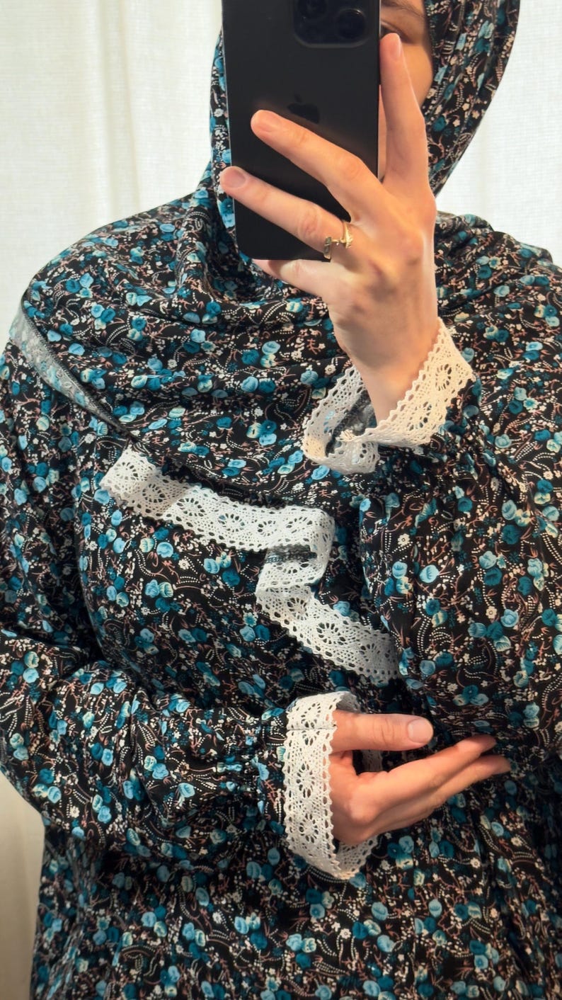 May include: A black garment with a teal floral pattern and white lace trim. The garment has a hood and white lace detailing on the sleeves and neckline. The fabric is patterned with small teal flowers and white accents.