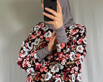 May include: A long-sleeved floral print dress with a black background, featuring red, white, and brown flowers. The dress is paired with a gray hijab and long-sleeved undershirt. The person is holding a black phone.
