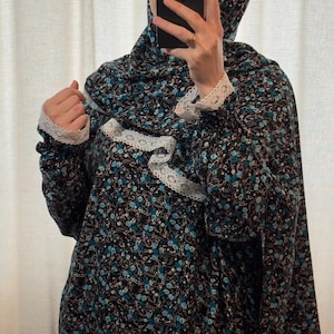 May include: A black prayer dress and headscarf set with a blue floral design and white lace trim. The dress features long sleeves with lace cuffs. The headscarf matches the dress's floral pattern. The outfit is suitable for prayer.