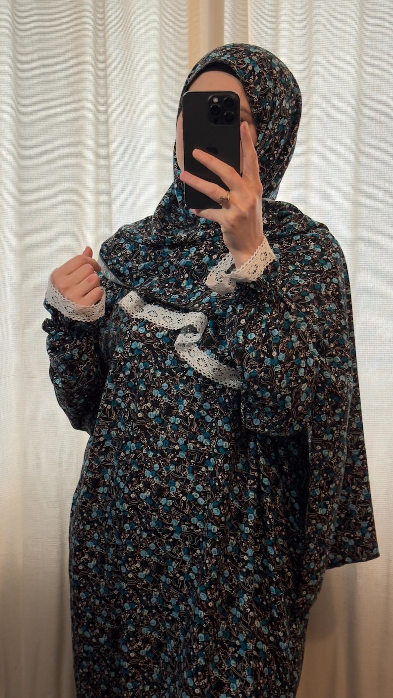May include: A black prayer dress and headscarf set with a blue floral design and white lace trim. The dress features long sleeves with lace cuffs. The headscarf matches the dress's floral pattern. The outfit is suitable for prayer.