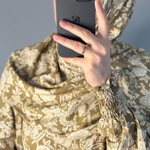 May include: Olive green floral print hijab and matching top. The fabric features a repeating pattern of white flowers and leaves. The top has gathered sleeves. A black phone is held in front of the face.