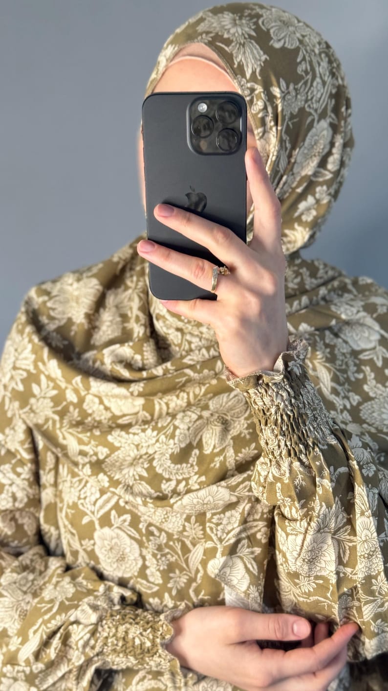 May include: Olive green floral print hijab and matching top. The fabric features a repeating pattern of white flowers and leaves. The top has gathered sleeves. A black phone is held in front of the face.