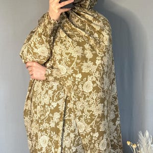 May include: An olive green floral print outfit, including a head covering and long-sleeved garment. The fabric features a detailed white floral pattern. The outfit is worn by a person holding a phone. The background is a solid grey wall.