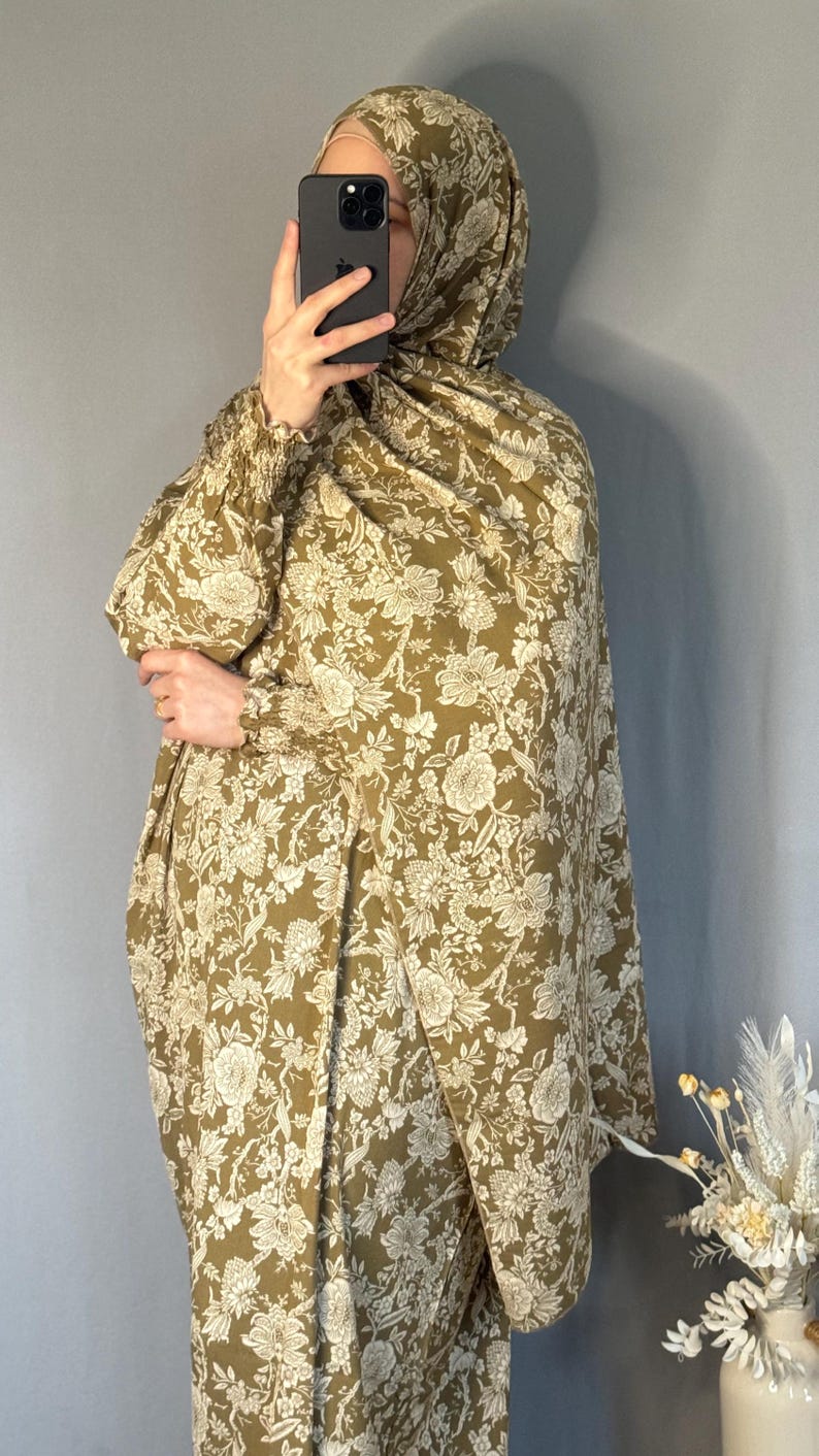 May include: An olive green floral print outfit, including a head covering and long-sleeved garment. The fabric features a detailed white floral pattern. The outfit is worn by a person holding a phone. The background is a solid grey wall.