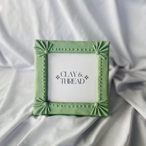 May include: A square, light green picture frame with decorative embellishments. The frame has a series of small, round dots along the edges and stylized floral designs at each corner. The text "CLAY & THREAD" is visible within the frame.