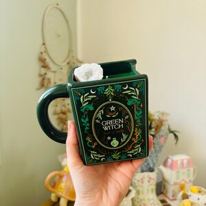 Peut inclure: Un mug en céramique vert foncé en forme de livre rectangulaire avec une anse arrondie. L'avant présente des illustrations botaniques dorées et blanches, un croissant de lune et le texte "GREEN WITCH Book of Spells". Un cristal blanc est à l'intérieur.
