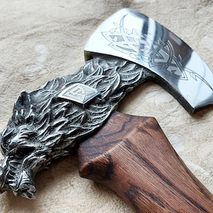 May include: A hand-forged axe with a wolf head design on the handle and a Celtic knot design on the blade. The axe has a wooden handle with a dark brown stain.