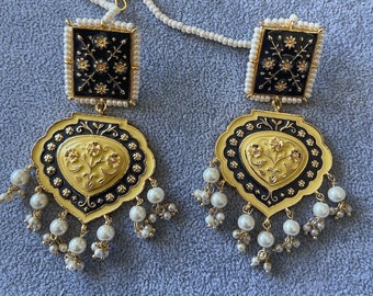 May include: A pair of gold earrings with black enamel and pearl accents. The earrings feature a square top with floral designs and a heart-shaped bottom with floral designs. The earrings are connected by a pearl chain.