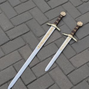 Könnte beinhalten: Zwei Metallschwerter mit silberfarbenen Klingen, goldfarbenen Parierstangen und braun lederumwickelten Griffen. Jedes Schwert hat einen goldfarbenen Knauf und eine goldfarbene Platte nahe dem Heft mit dunklen, runenartigen Gravuren.