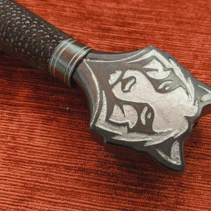 May include: Close-up of a sword hilt with a textured, dark brown grip and a silver-coloured guard. The guard features an engraved design with floral and animal motifs. The sword rests on a textured, reddish-orange surface.