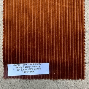 Puede incluir: Un primer plano de una tela de pana de color óxido con una etiqueta que dice "IMPORTED 6 WALE CORD Rusty 6 Wale Corduroy 57" 8.5 oz 100% Cotton 1,396 Yards".