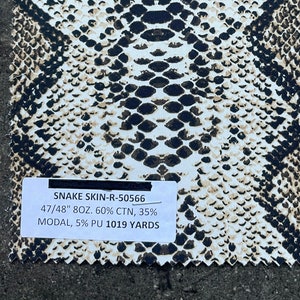 May include: A close-up of a white fabric with a black and brown snake skin pattern. The fabric is labeled "SNAKE SKIN-R-50566 47/48" 8OZ. 60% CTN, 35% MODAL, 5% PU 1019 YARDS".