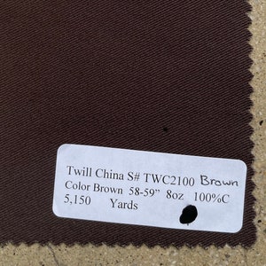 Op de afbeelding: Een vierkant staal van bruine keperstof met een getextureerd oppervlak. Een wit label toont de tekst "Twill China S# TWC2100 Brown Color Brown 58-59" 8oz 100%C 5,150 Yards". De stof is ongeveer 147-150 cm breed.
