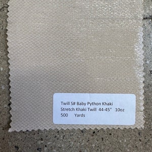 May include: A beige fabric sample with a textured finish. A white label reads "Twill S# Baby Python Khaki Stretch Khaki Twill 112-114cm 10oz 500 Yards". The fabric is displayed on a grey concrete surface.