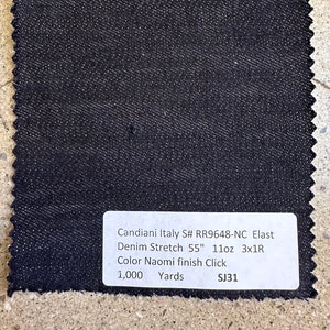 May include: A square swatch of dark blue denim fabric with a textured surface. The fabric has a raw, jagged edge. A white label displays text: "Candiani Italy S# RR9648-NC Elast Denim Stretch 55" 11oz 3x1R Color Naomi finish Click 1,000 Yards SJ31".