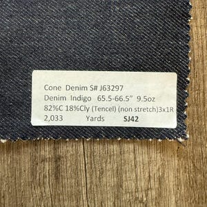 May include: A swatch of dark indigo denim fabric with a white label. The label reads "Cone Denim S# J63297" and lists fabric details including 65.5-66.5" width, 9.5oz weight, and fiber composition. The fabric is non-stretch.