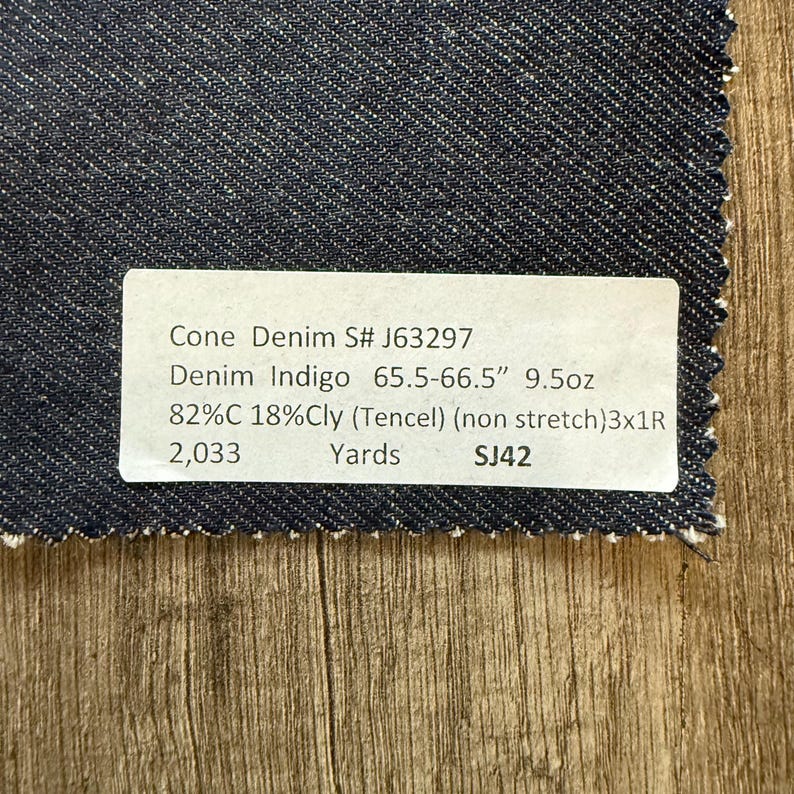 May include: A swatch of dark indigo denim fabric with a white label. The label reads "Cone Denim S# J63297" and lists fabric details including 65.5-66.5" width, 9.5oz weight, and fiber composition. The fabric is non-stretch.