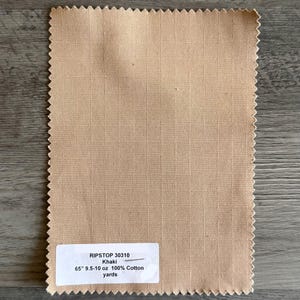 May include: A sample of khaki-colored Ripstop 30310 fabric. The fabric has a grid pattern and a raw, zig-zag edge. The label indicates it is 65 inches wide and made of 100% cotton.