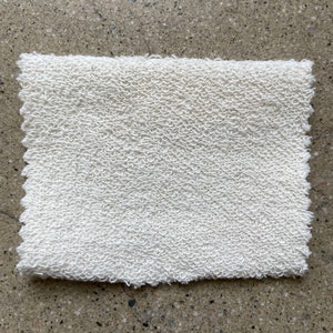 May include: A white, textured fabric swatch. The fabric has a slightly rough, uneven surface.