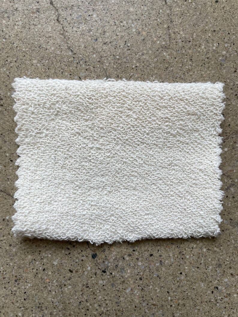 May include: A white, textured fabric swatch. The fabric has a slightly rough, uneven surface.