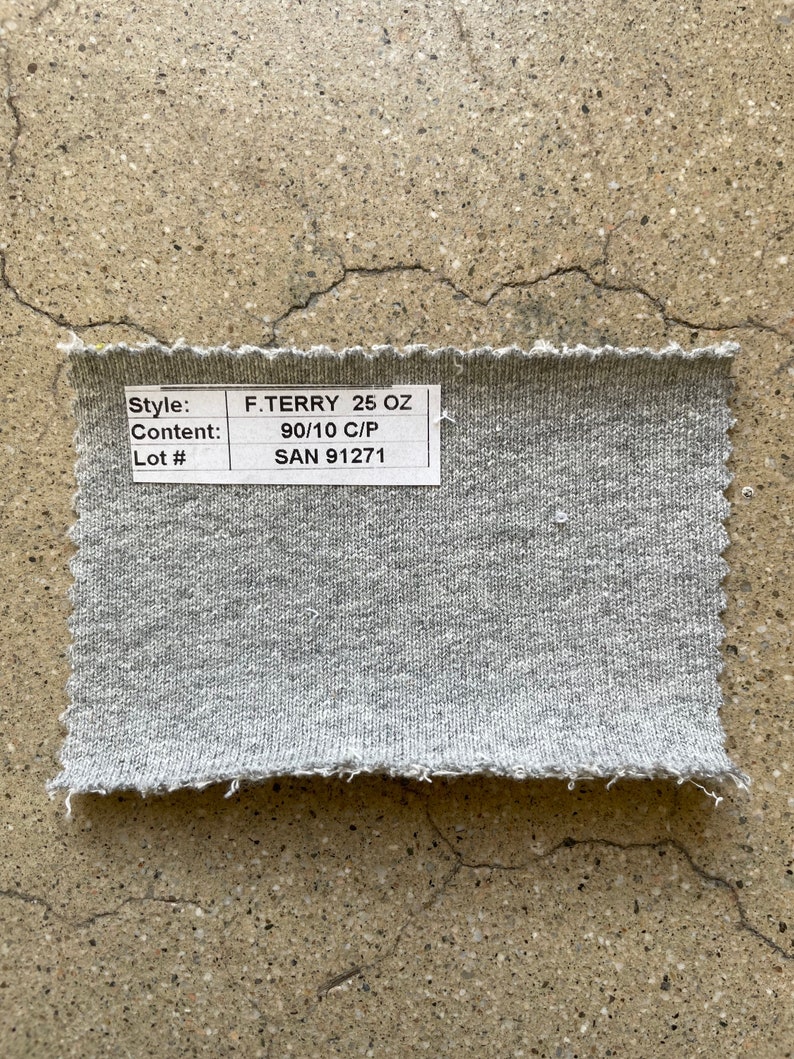 May include: A close-up of a gray fabric swatch with a white label that reads "Style: F.TERRY 25 OZ", "Content: 90/10 C/P", and "Lot #: SAN 91271".