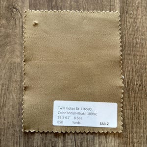 Op de afbeelding: Een staal van kaki-kleurige twill stof met een zigzagrand. De stof heeft een diagonaal weefpatroon. Een wit label toont de tekst: "Twill Indian S# 116580 Color British-Khaki 100%C 151-155cm 8.5oz 650 Yards SA3-2."
