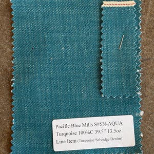 May include: A sample of turquoise denim fabric with a selvedge edge. The fabric is labeled "Pacific Blue Mills S#SN-AQUA Turquoise 100%C 39.5" 13.5oz Line Item (Turquoise Selvedge Denim)". The fabric has a textured appearance and a teal color.