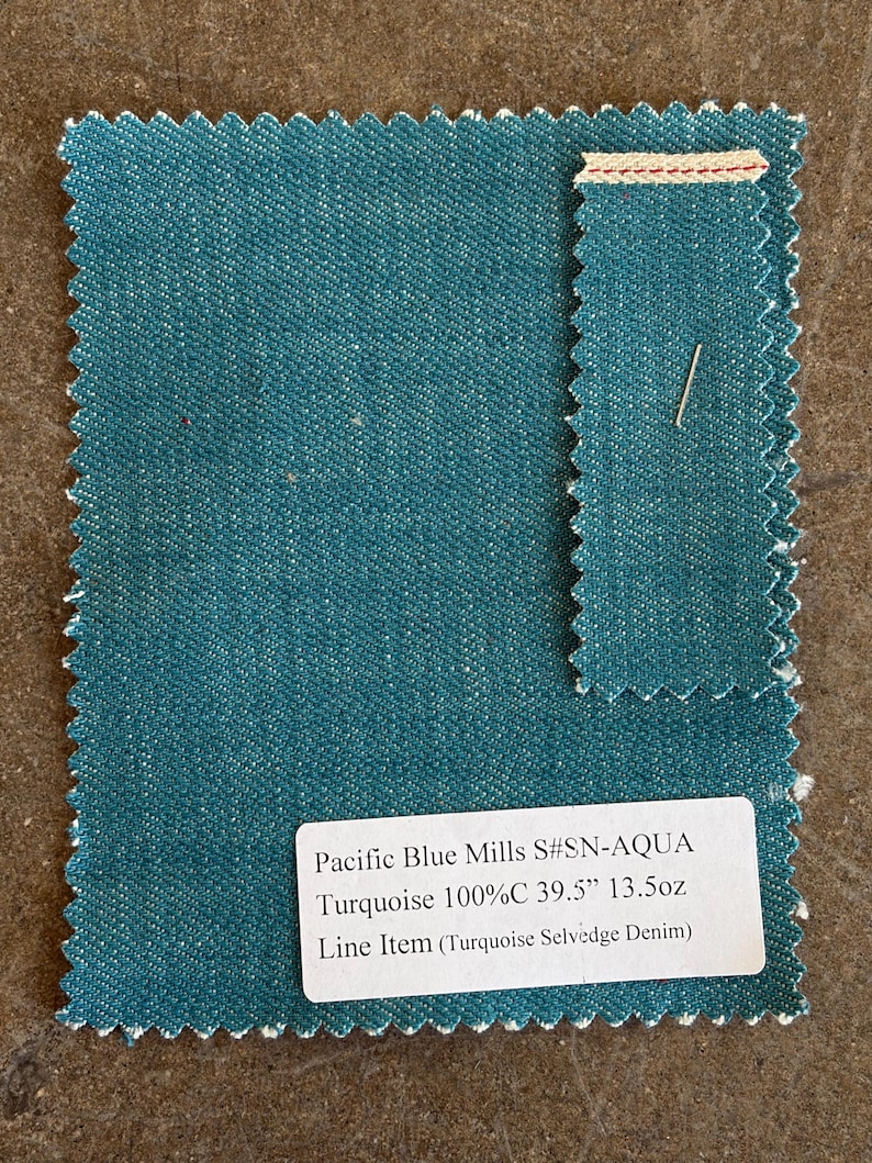 May include: A sample of turquoise denim fabric with a selvedge edge. The fabric is labeled "Pacific Blue Mills S#SN-AQUA Turquoise 100%C 39.5" 13.5oz Line Item (Turquoise Selvedge Denim)". The fabric has a textured appearance and a teal color.