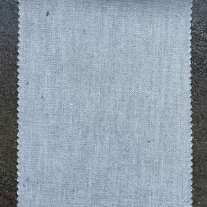 May include: A close-up of a white fabric with a subtle diagonal weave pattern. The fabric has a slightly rough texture and a slightly uneven edge.