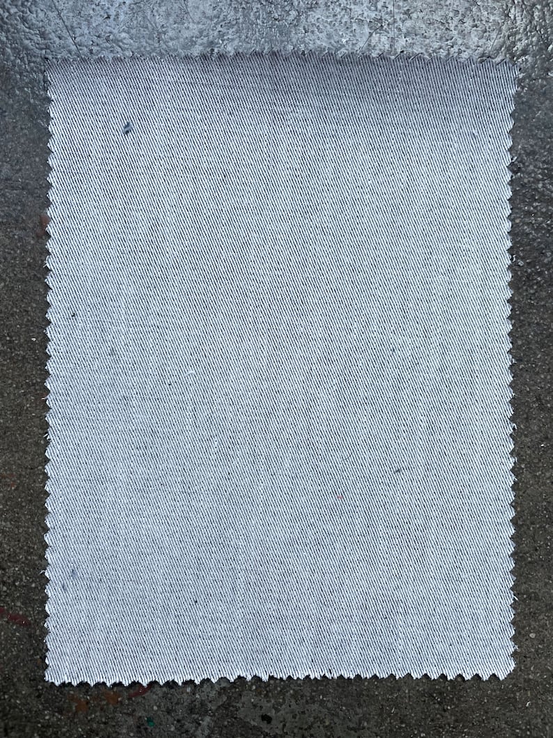 May include: A close-up of a white fabric with a subtle diagonal weave pattern. The fabric has a slightly rough texture and a slightly uneven edge.