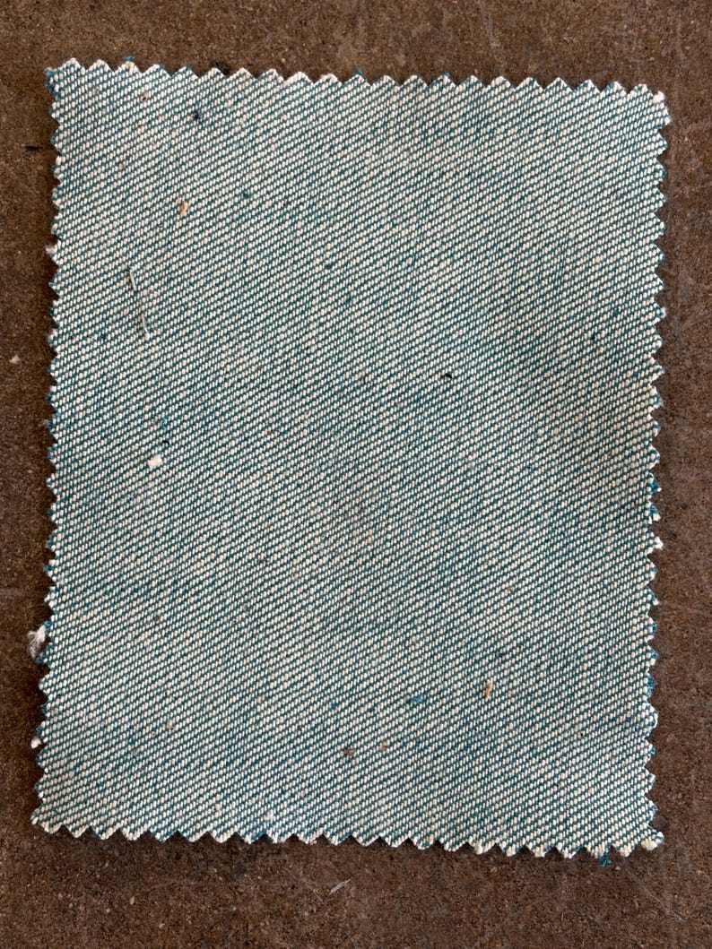 May include: A square fabric swatch with a teal and white diagonal weave pattern. The edges are cut with a zig-zag pattern. The fabric has a textured appearance with small flecks of color throughout. The swatch is likely for textiles or crafting.