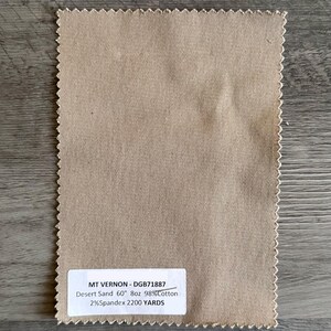 May include: A rectangular swatch of Desert Sand fabric with a serged edge. The fabric has a diagonal weave pattern. A white label displays the text "MT VERNON - DGB71887 Desert Sand 60" 8oz 98%Cotton 2%Spandex 2200 YARDS."