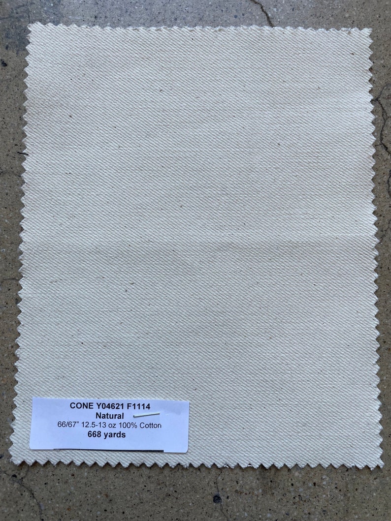 Puede incluir: Un primer plano de una muestra de tela blanca natural con una etiqueta que dice "CONE Y04621 F1114 Natural 66/67" 12.5-13 oz 100% Algod&oacute;n 668 yardas"