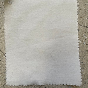 May include: A close-up of a plain, off-white fabric swatch. The fabric has a slightly textured surface and a slightly uneven edge.