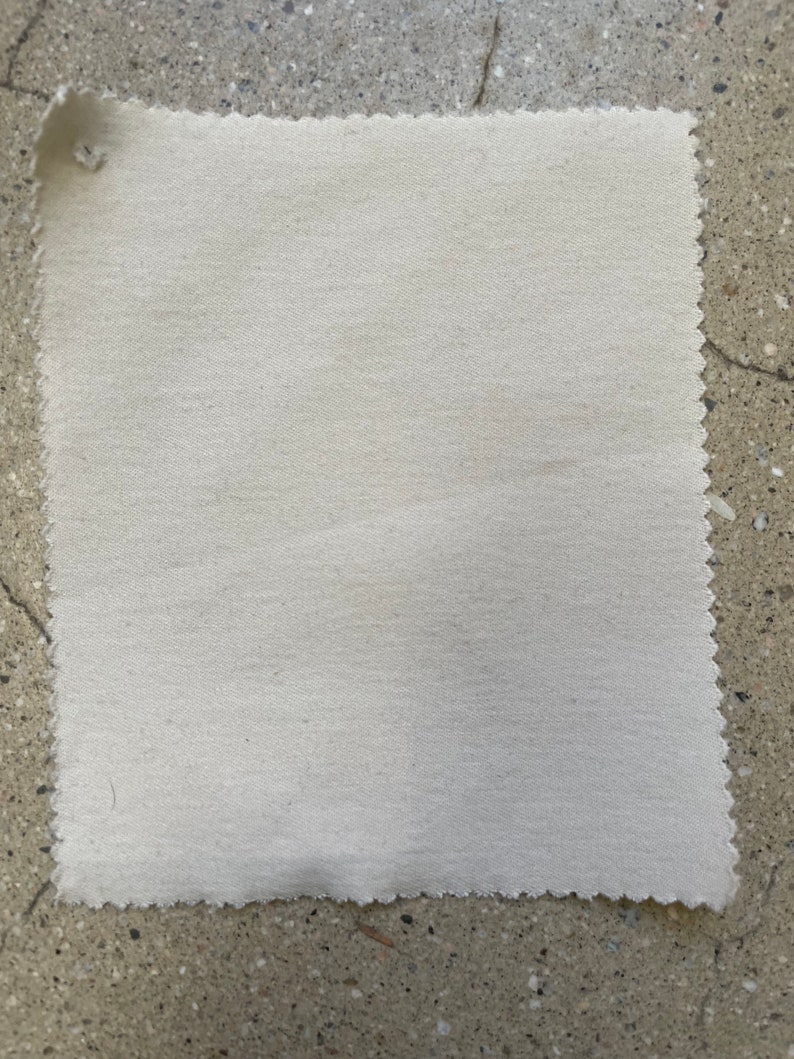 May include: A close-up of a plain, off-white fabric swatch. The fabric has a slightly textured surface and a slightly uneven edge.