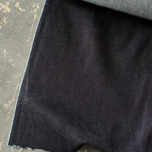 May include: A roll of dark indigo denim fabric. The fabric is a deep, saturated blue with a subtle texture. The edge of the fabric is visible, showing the lighter blue underside. The fabric is unrolled on a concrete surface.
