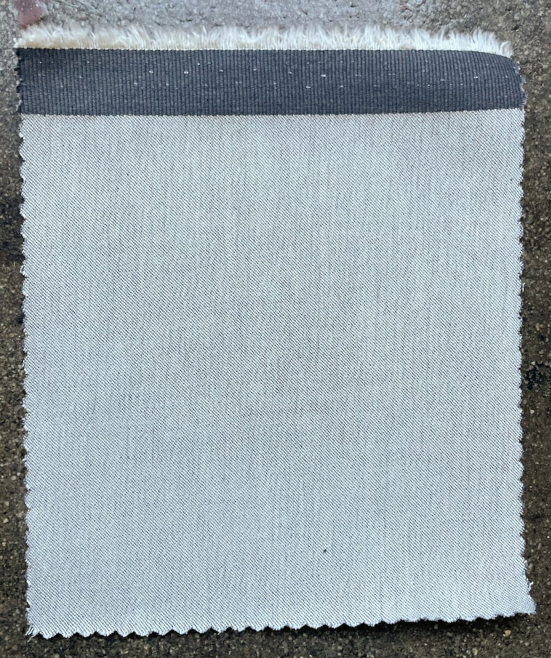 May include: A close-up of a light gray fabric with a black woven trim. The fabric has a subtle diagonal weave pattern.