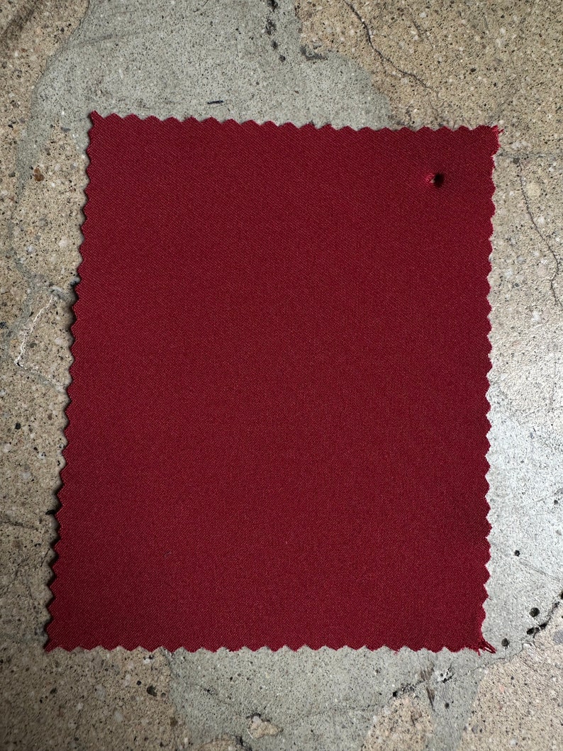 May include: A close-up of a solid burgundy fabric swatch. The fabric has a subtle texture and a slightly scalloped edge.