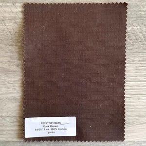 May include: A dark brown fabric swatch with a grid pattern. The fabric has a textured appearance and a raw, jagged edge. A white label at the bottom displays the text "RIPSTOP 26679 Dark Brown 64/65" 7 oz 100% Cotton yards."