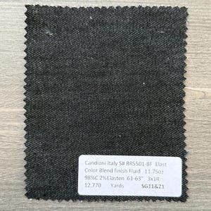 May include: A dark denim fabric swatch with a textured surface. The swatch has a jagged edge and a white label with text that reads "Candiani Italy S# RR5501-BF Elast Color Blend finish Fluid 11.75oz 98%C 2% Elasten 61-63" 3x1R 12,770 Yards SG11&21."