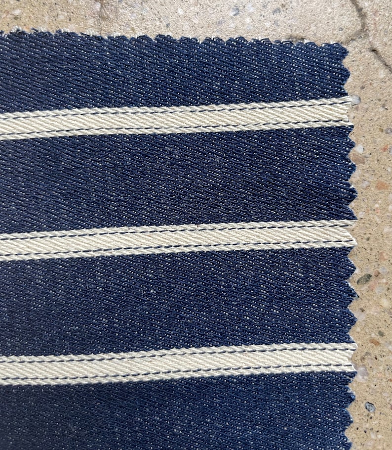Puede incluir: Primer plano de una tela azul marino con rayas blancas. La tela tiene una superficie texturizada y un borde ligeramente irregular.