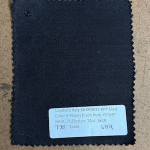 May include: A square swatch of dark navy blue fabric with a serged edge. A white label with black text provides details about the fabric, including the manufacturer, color, and composition. The fabric appears to be a medium weight material.
