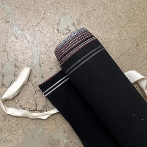 Puede incluir: Tela negra enrollada con un borde a rayas en rojo, blanco y gris. Se ve una cinta de tela blanca. La tela parece ser un material pesado, posiblemente para ropa o decoración del hogar.