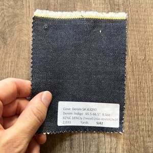 May include: A swatch of dark indigo denim fabric with a raw edge and a yellow stripe. The fabric sample has a label with details including "Cone Denim S# J63297" and "Denim Indigo 65.5-66.5" 9.5oz".
