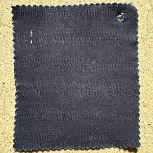 May include: A dark gray fabric swatch with a scalloped edge. The fabric has a woven texture and a small hole in the upper right corner.