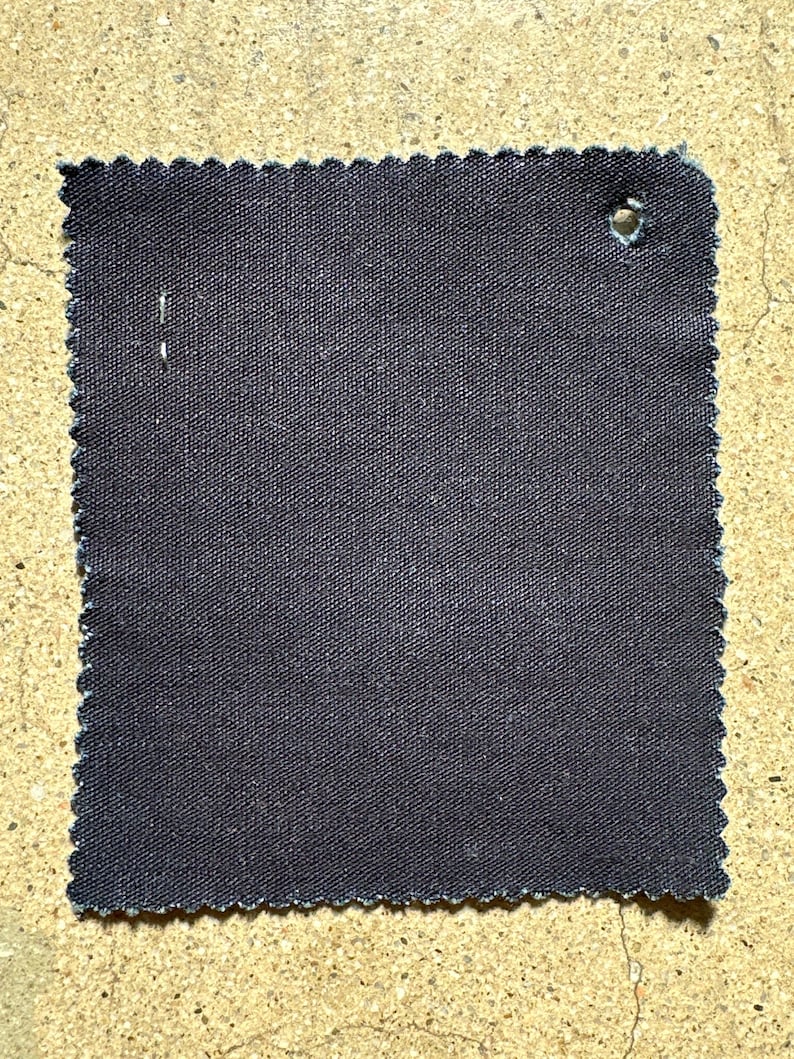 May include: A dark gray fabric swatch with a scalloped edge. The fabric has a woven texture and a small hole in the upper right corner.
